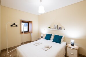 Una camera d'albergo con un letto, comodini, un appendiabiti, una piccola finestra e uno scaffale a parete con delle foto.