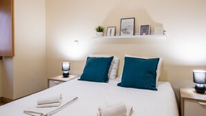 A neatly made bed with two pillows, a bedside table with a lamp, and a shelf with framed pictures and a plant.