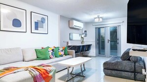 Ein modernes Wohnzimmer mit einer Couch, einem Couchtisch und einem grauen Sofa. Es gibt einen Essbereich mit Tisch und Stühlen sowie eine Küche mit Kühlschrank und Mikrowelle.