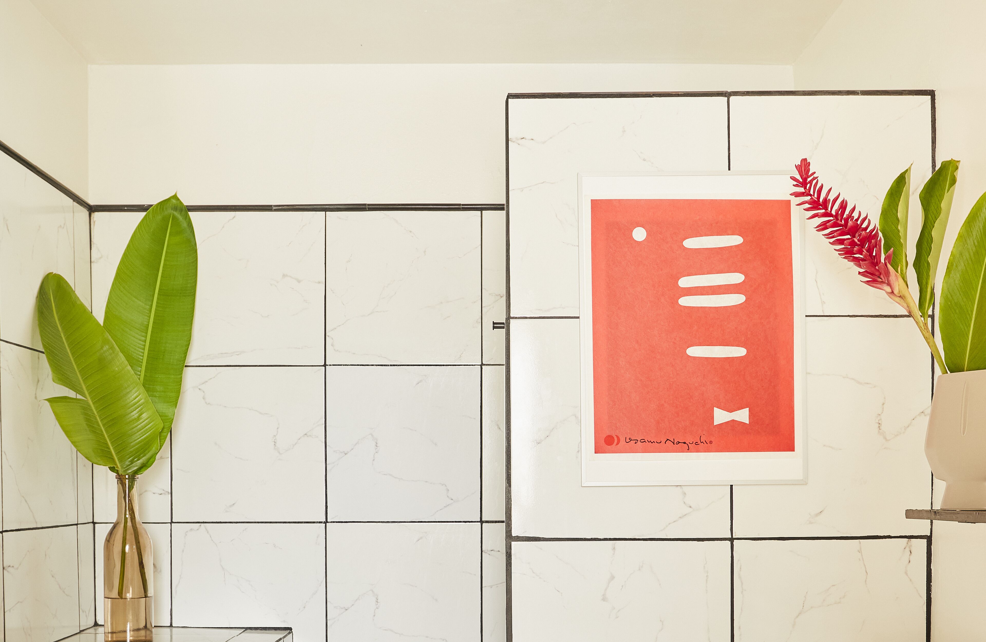 A bathroom with white marble tiles, a framed artwork, and two potted plants.