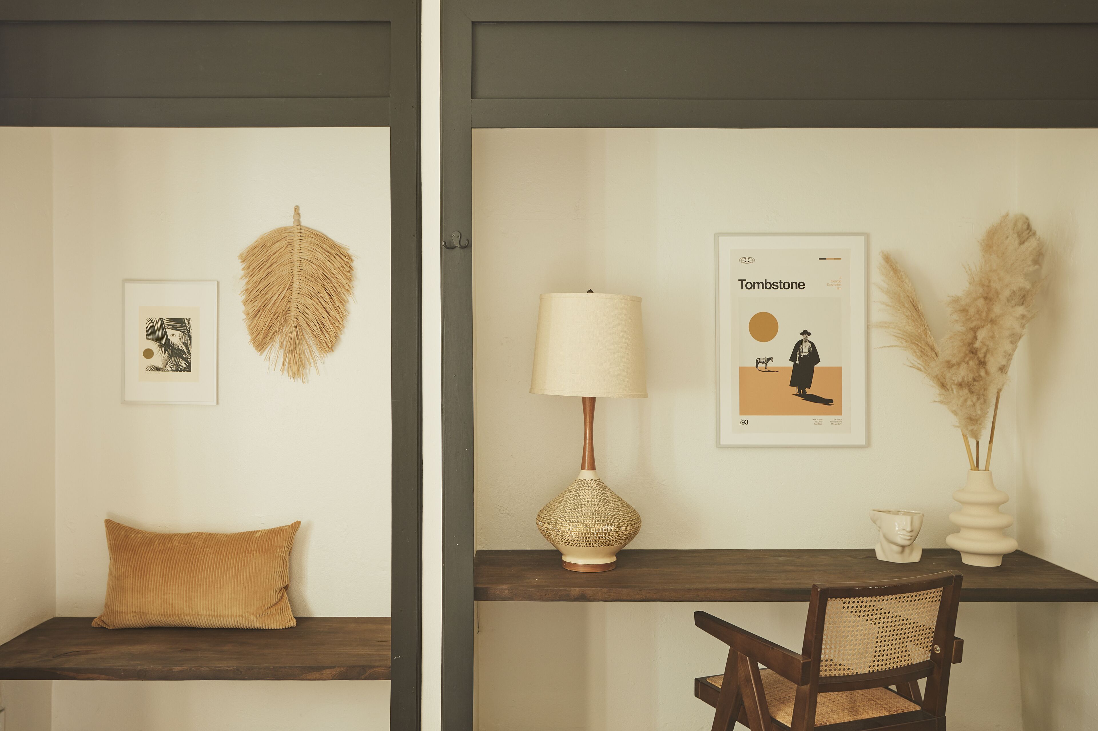 A room with a wooden desk, a chair, a lamp, a framed picture, a decorative leaf, and a vase with pampas grass.