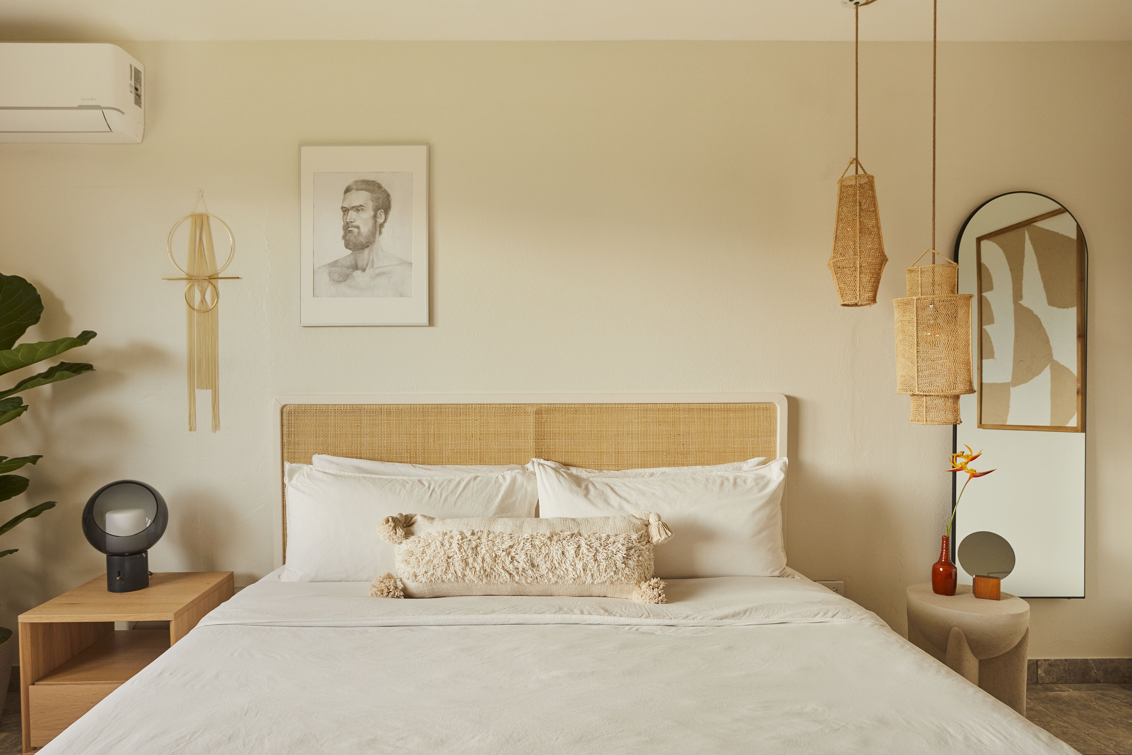 A neatly made bed with white linens, a headboard with a woven texture, a bedside table with a lamp, and a wall-mounted mirror.