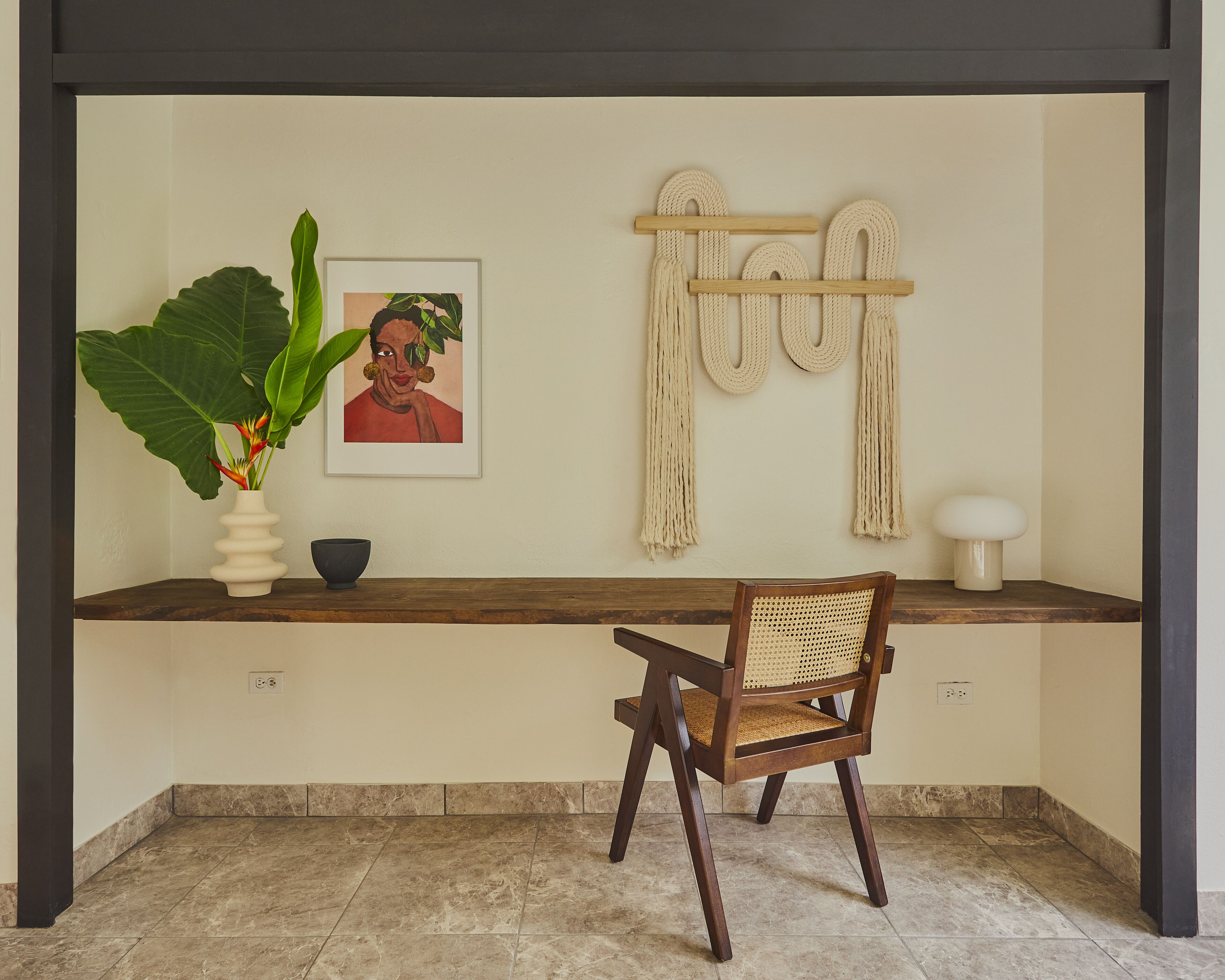 A room with a wooden desk, a chair, a potted plant, a framed picture, macramé wall hangings, and a white lamp.