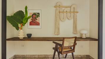 A room with a wooden desk, a chair, a potted plant, a framed picture, macramé wall hangings, and a white lamp.