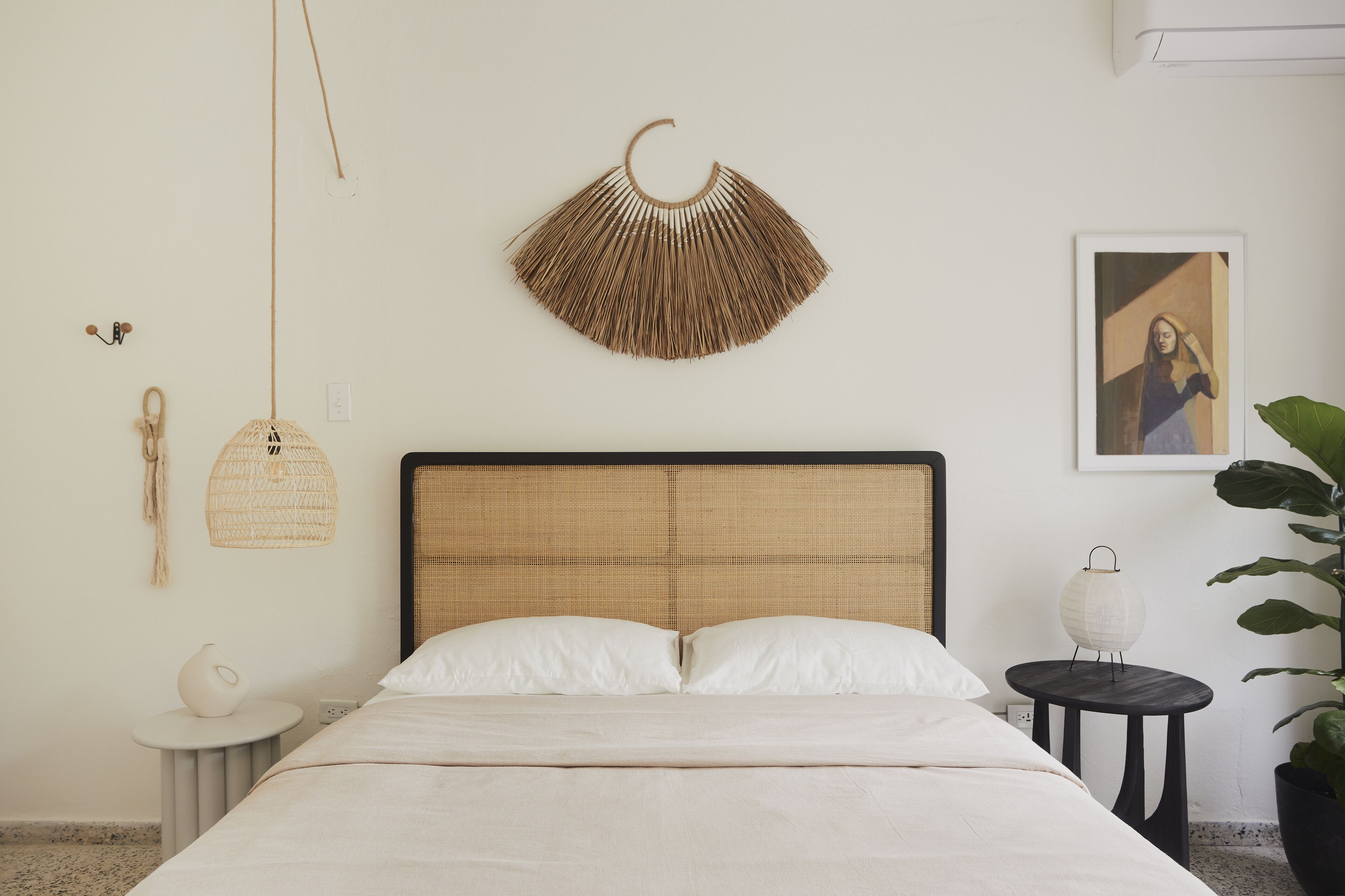 A bedroom with a bed, a hanging light, a wall-mounted fan, a small table with a lamp, and a framed picture on the wall.