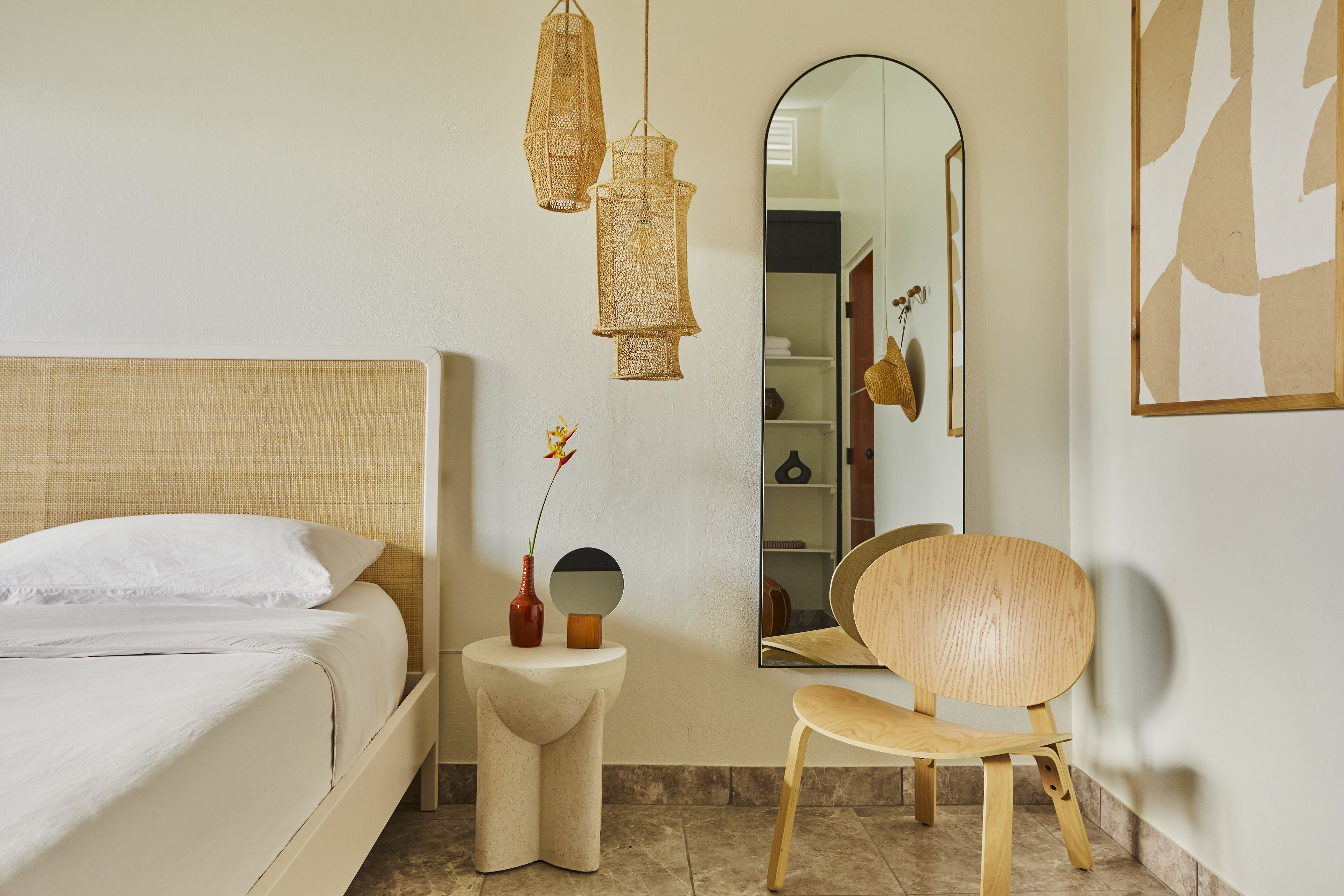 A bedroom with a bed, a small side table with a vase, a chair, and a mirror on the wall.