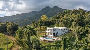 Ein modernes, weißes Haus mit Pool, umgeben von üppiger Vegetation und Bergen.