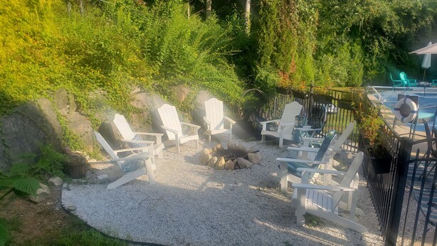 Un patio con sillas Adirondack blancas dispuestas alrededor de una fogata, rodeado de grava y follaje.