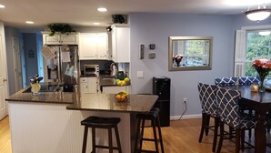 Una cocina moderna con gabinetes blancos, encimera de granito, horno integrado y refrigerador. Hay dos taburetes de bar y una mesa de comedor con cojines estampados.