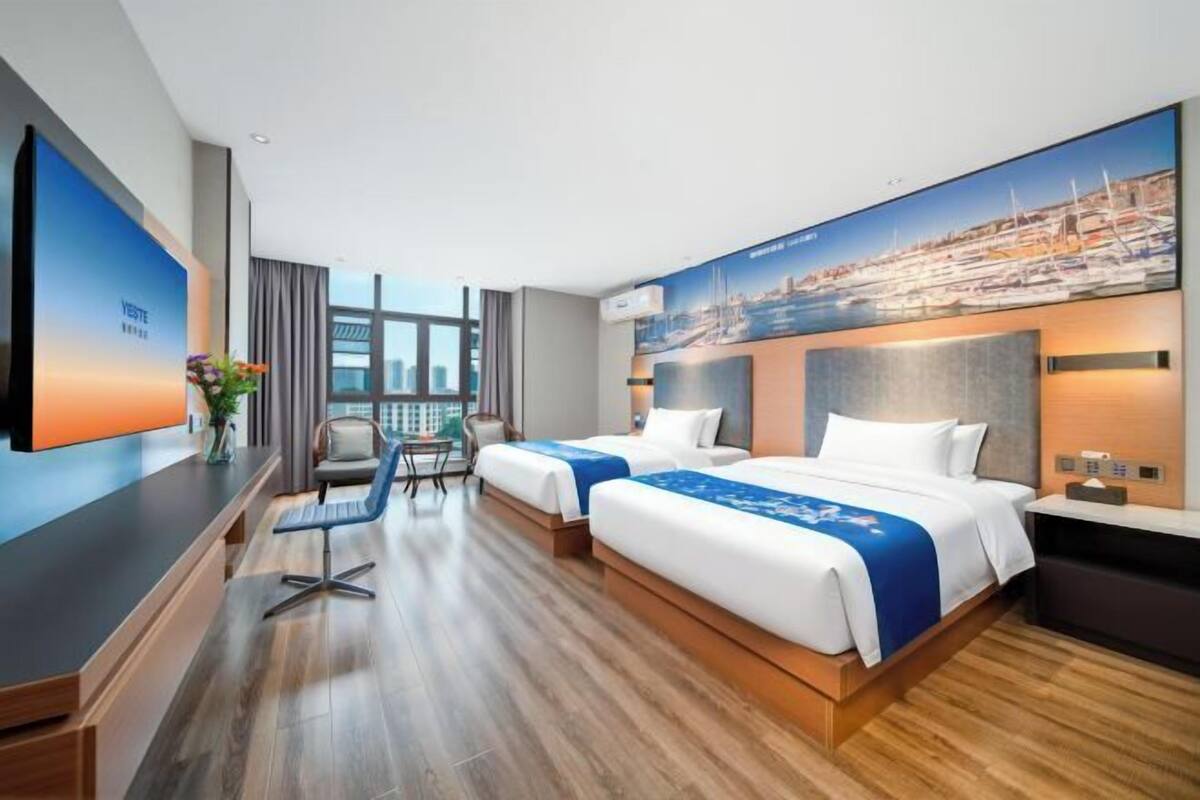 A hotel room with two beds, a desk, a TV, and a cityscape mural.