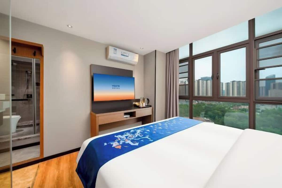 A hotel room with a bed, a TV, a shower, and a view of buildings.