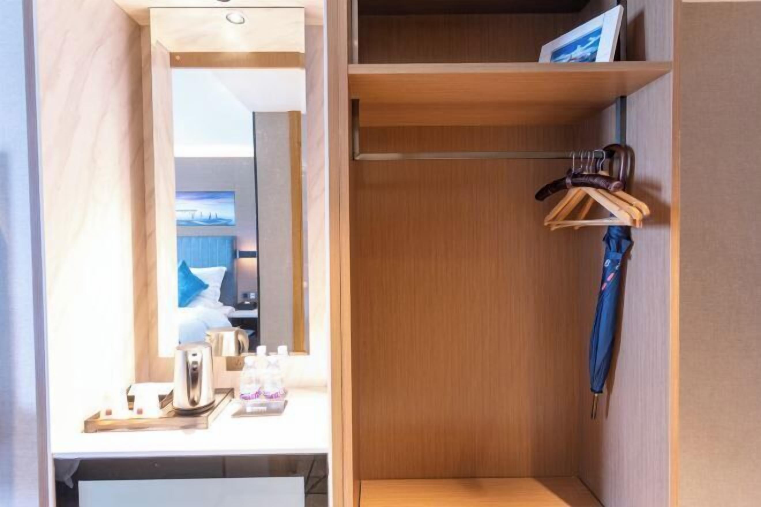 A hotel room with a wooden vanity, a mirror, a small table with a kettle and bottles, and a hanging blue umbrella.