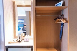 Quarto de hotel com uma penteadeira de madeira, um espelho, uma mesinha com chaleira e garrafas, e um guarda-chuva azul pendurado.