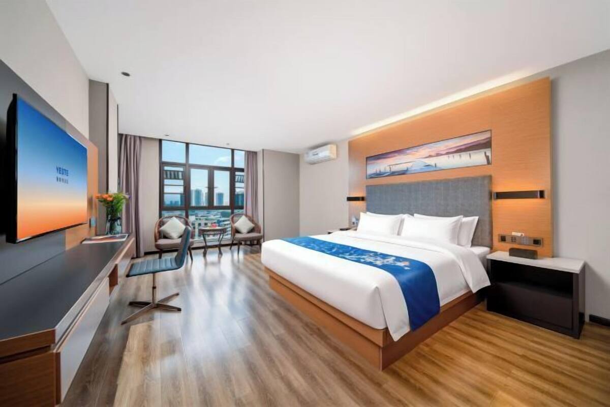 A modern hotel room with a large bed, a desk, a chair, a TV, and a city view.