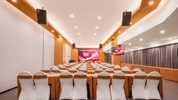 A conference room with rows of tables and chairs, a large screen at the front, and speakers on the ceiling.