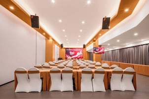 Un salón de conferencias con filas de mesas y sillas, una pantalla grande al frente y altavoces en el techo.