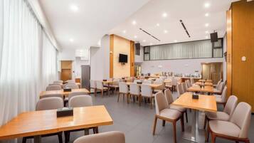 Un espace repas moderne avec plusieurs tables et chaises, une cuisine en arrière-plan et une grande fenêtre sur la gauche.