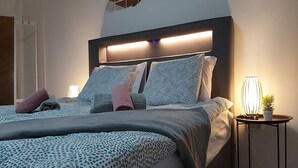 Une chambre à coucher moderne avec un grand lit, une literie à motifs, une tête de lit dotée d’un éclairage intégré et une table de chevet avec une lampe et une plante.