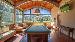 Una mesa de billar en una habitación con techo de madera y ventanas amplias, mobiliario de mimbre y un cuadro en la pared.