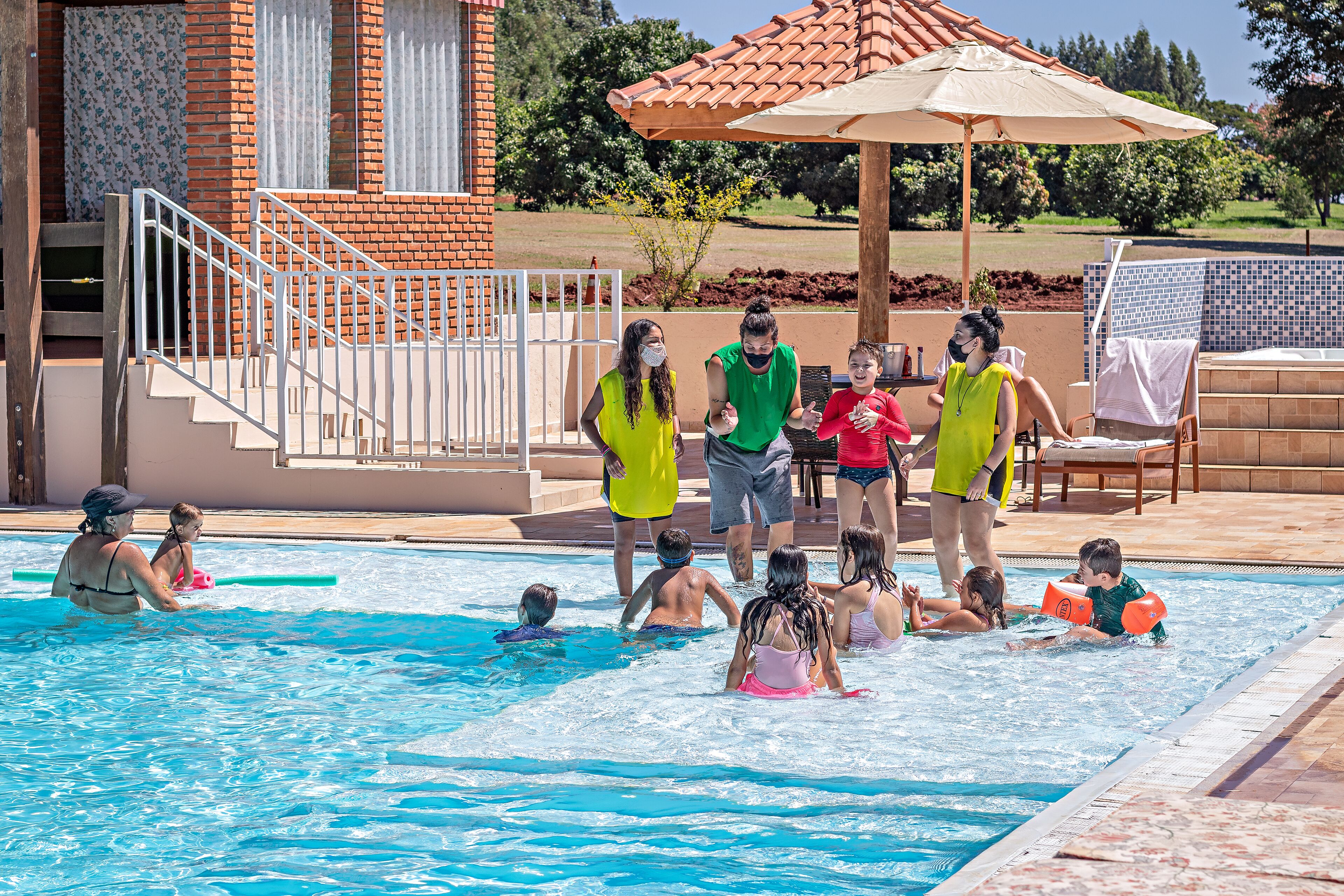 A swimming pool area with a brick building, a covered seating area, and a tiled deck.