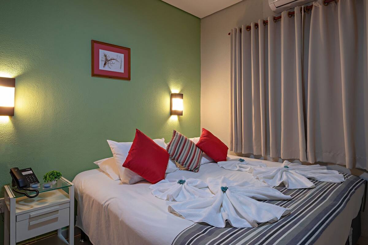 A hotel room with a bed, pillows, a bedside table with a phone, a wall-mounted light, and a window with curtains.
