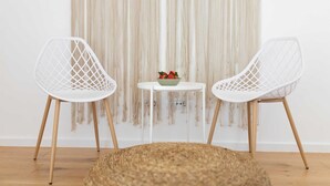 Dos sillas blancas con patas de madera, una mesa redonda con un cuenco de fruta y un tapete tejido sobre un piso de madera.
