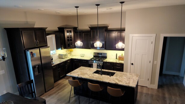 A modern kitchen with dark cabinets, a central island with bar stools, stainless steel appliances, and pendant lighting. - New Modern Home (Joplin)