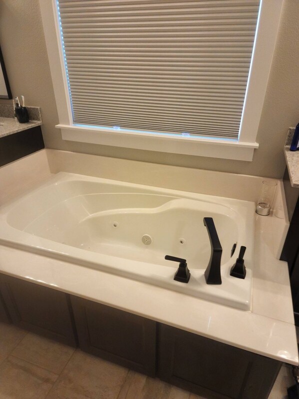 A modern bathroom with a large white bathtub, a window with blinds, and a small shelf with toiletries. - New Modern Home (Joplin)