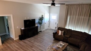 A living room with a flat-screen TV on a wooden stand, a brown sofa, a coffee table, and a ceiling fan. - New Modern Home (Joplin)