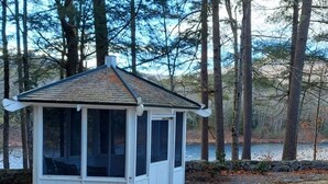 Un petit kiosque blanc, doté d’un toit en pente et de grandes fenêtres, entouré d’arbres et bordé par un plan d’eau.