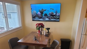 A small dining area with a wooden table, two chairs, a television mounted on the wall displaying an underwater scene, a potted plant, and a trash bin.