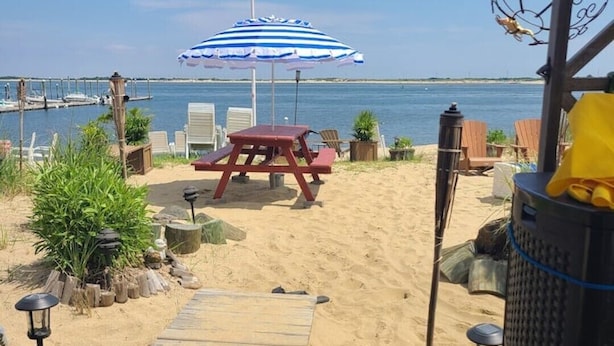 A wooden deck leading to a sandy beach area with a picnic table and chairs, a blue and white striped umbrella, and a view of the water.