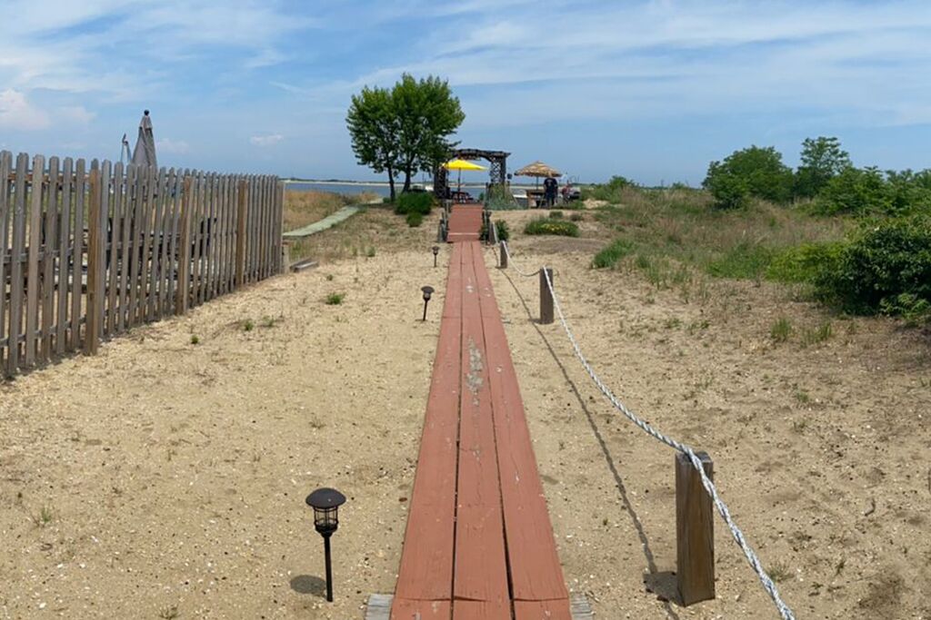 A wooden boardwalk leading to a beach area with a wooden fence on one side and a thatched structure with a yellow umbrella in the distance.