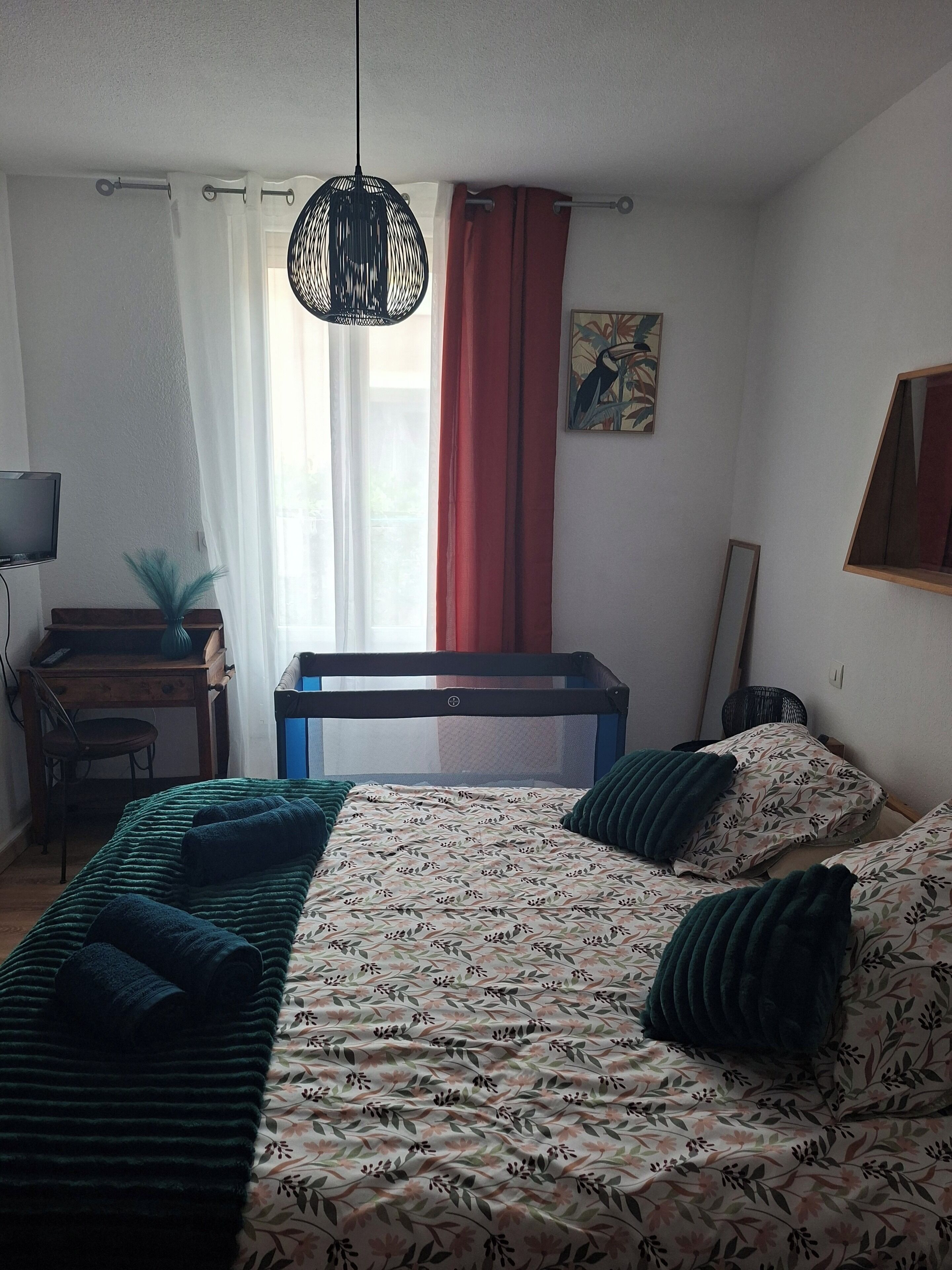 Une chambre à coucher comprenant un lit, un bureau, une chaise, un luminaire suspendu et une fenêtre avec des rideaux.