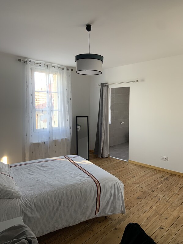 A bedroom with a bed, wooden flooring, a ceiling light, a window with curtains, and a door leading to a bathroom.