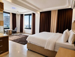 A modern hotel room with a large bed, a desk, a chair, and a view of the city.