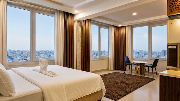 Habitación de hotel con cama, escritorio, dos sillas y ventanas amplias con vista a la ciudad.
