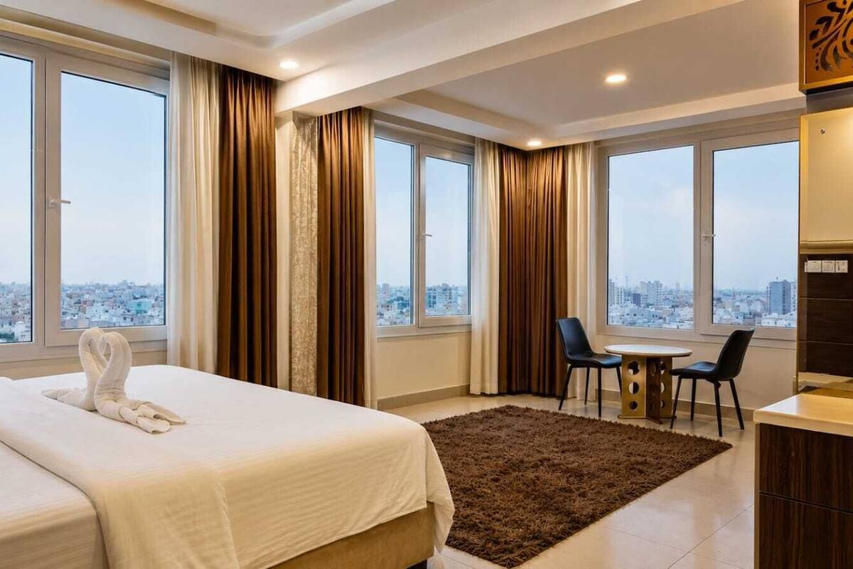 A hotel room with a bed, a desk, two chairs, and large windows with a city view.