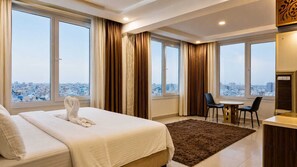A hotel room with a bed, a desk, two chairs, and large windows with a city view. - Diamond Star Hotel (Seeb)