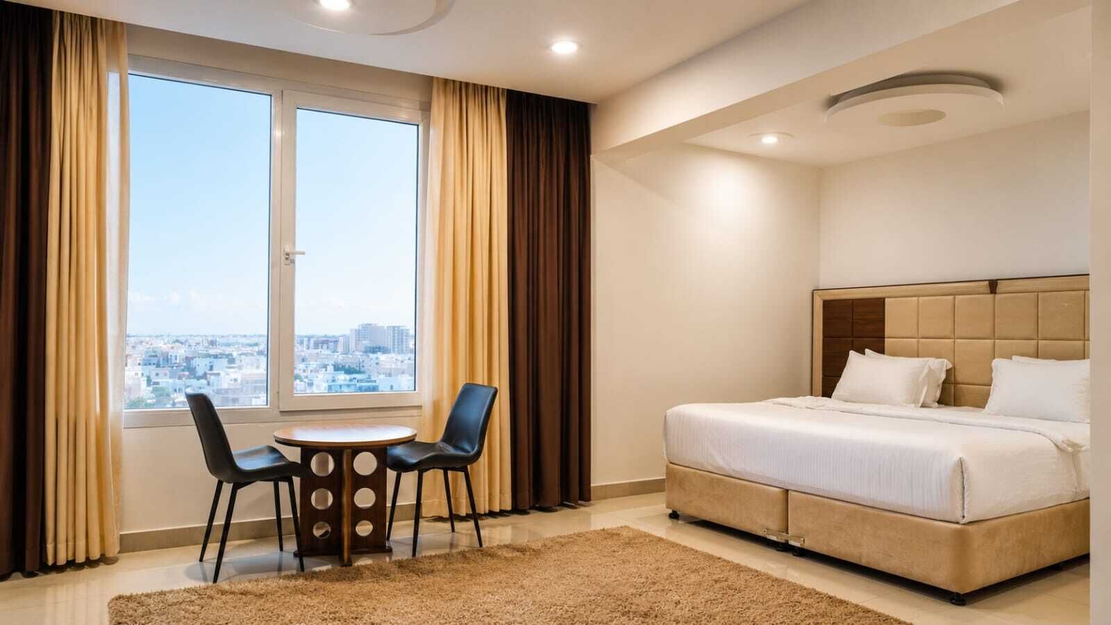 Habitación de hotel con cama, una pequeña mesa con sillas, vista de la ciudad a través de un ventanal grande y una lámpara en el techo.