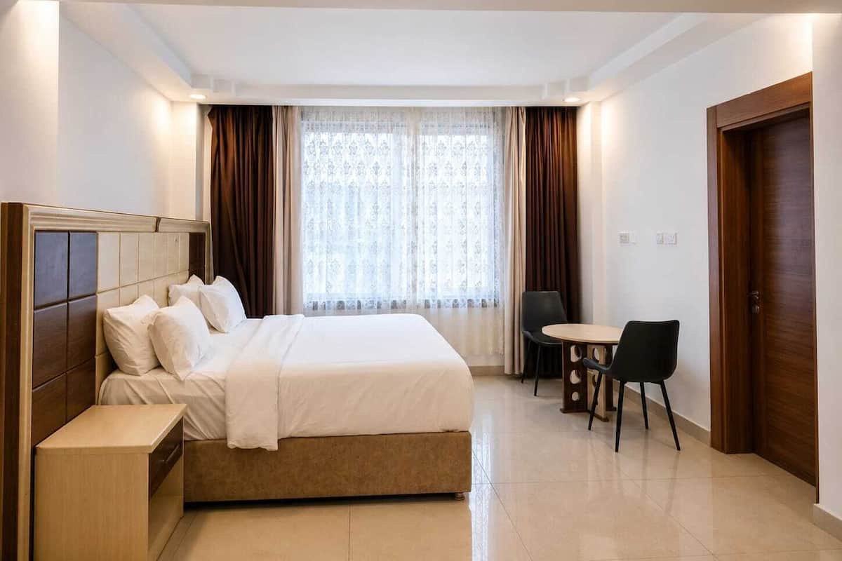A hotel room with a bed, bedside table, a small round table with two chairs, a window with curtains, and a door.