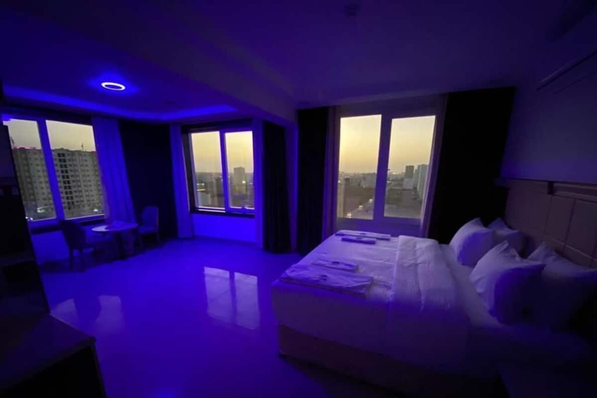 A hotel room with a large bed, a desk, and a view of the cityscape through the windows.