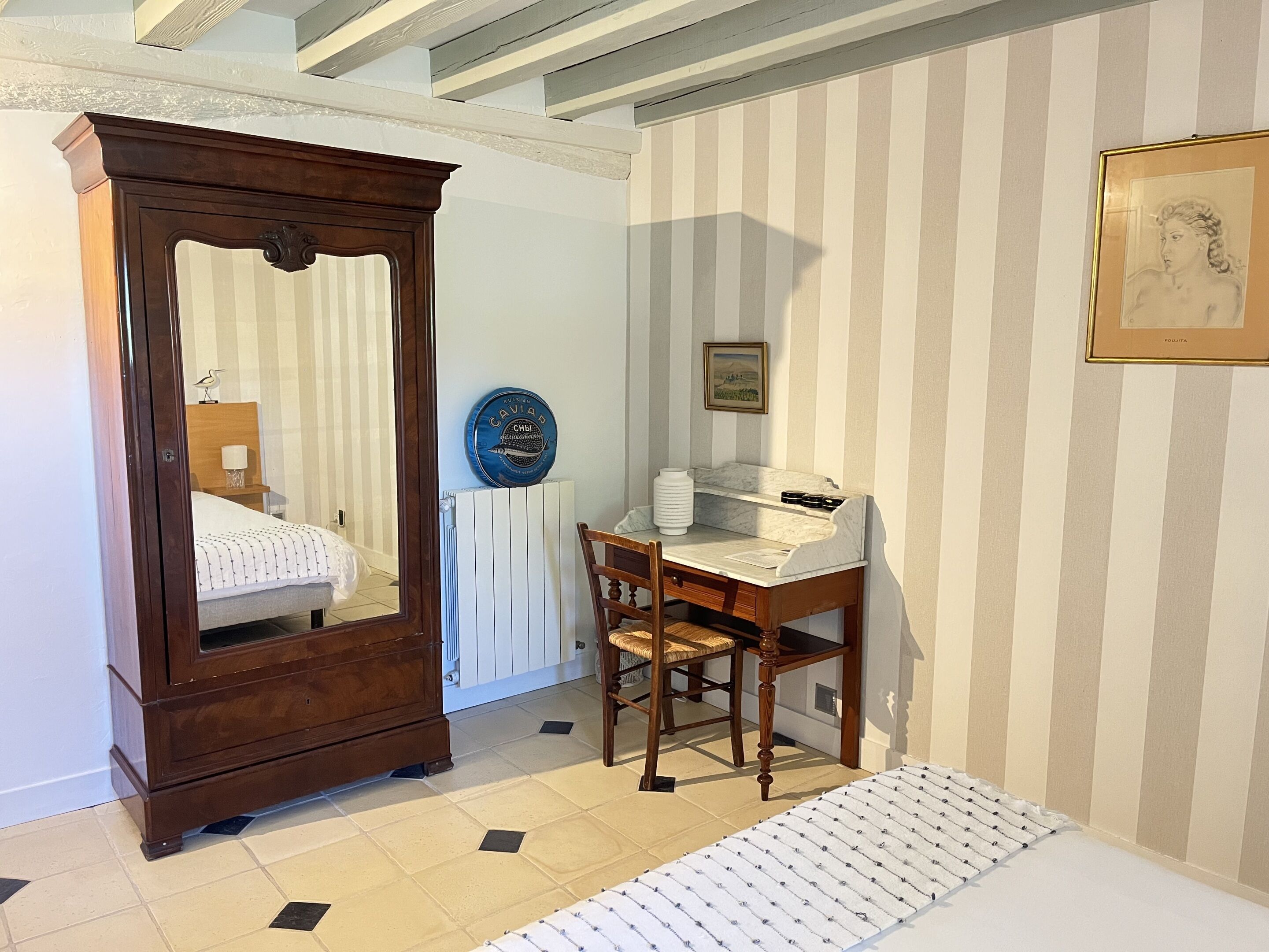 Una habitación con un armario de madera, una mesita de noche, una silla, un radiador, un cuadro enmarco y una pared a rayas.