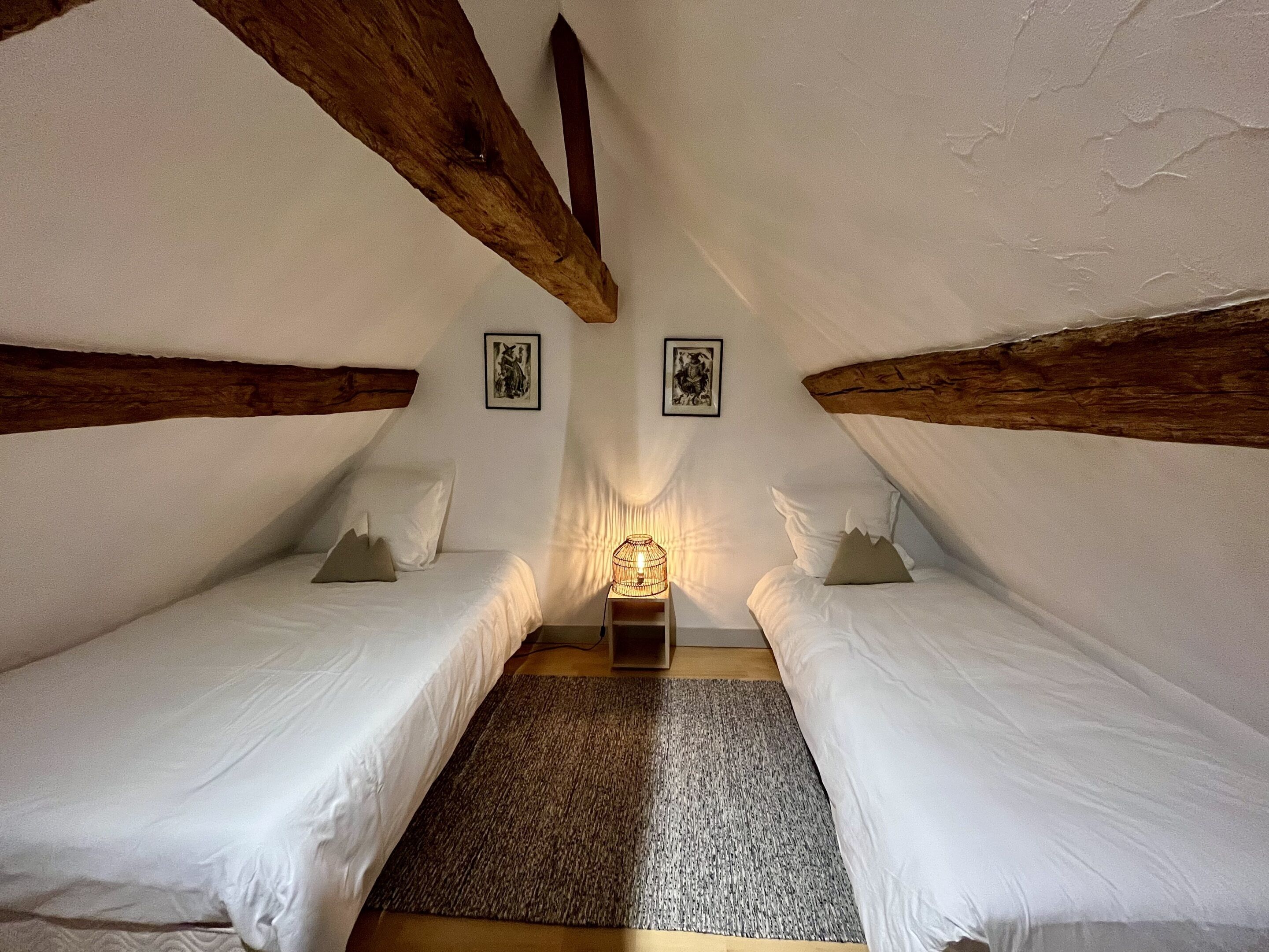 Un dormitorio acogedor en el ático, con dos camas individuales, techo con vigas de madera y una pequeña mesa con lámpara.
