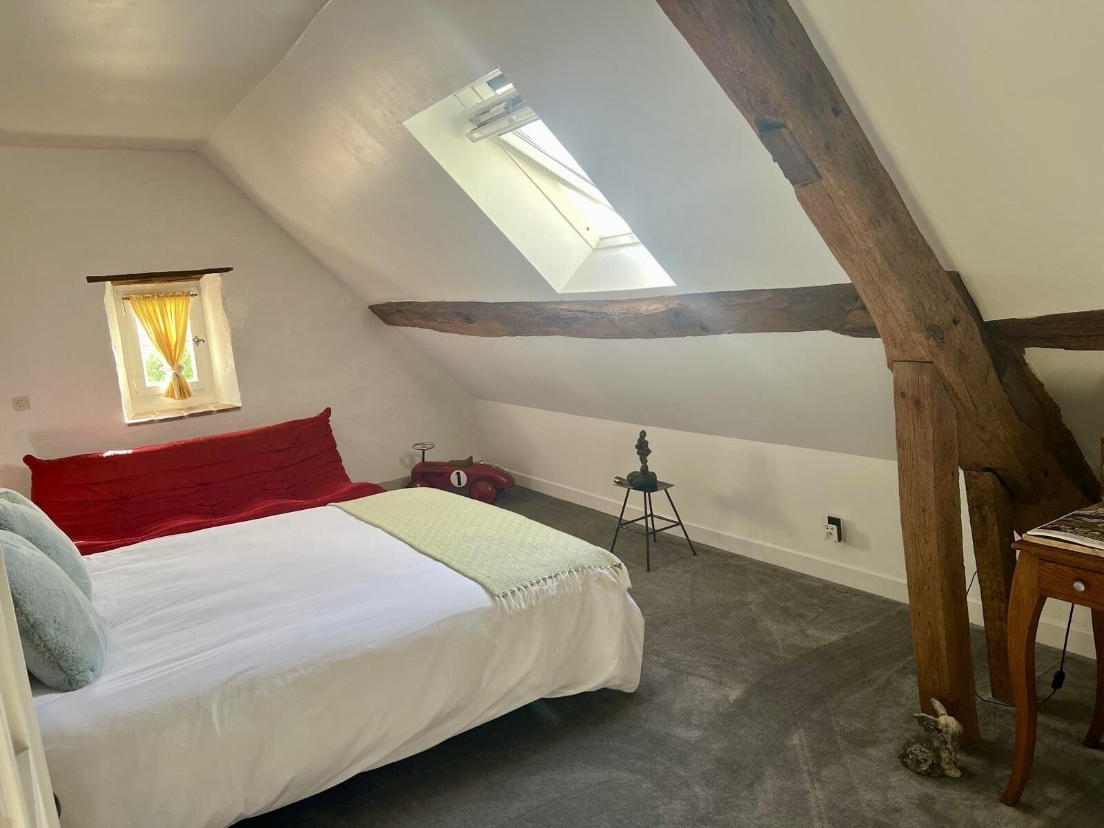 Een slaapkamer met een schuine plafond, een bed met rode en witte beddengoed, een klein tafeltje, een stoel en een raam met gordijnen.