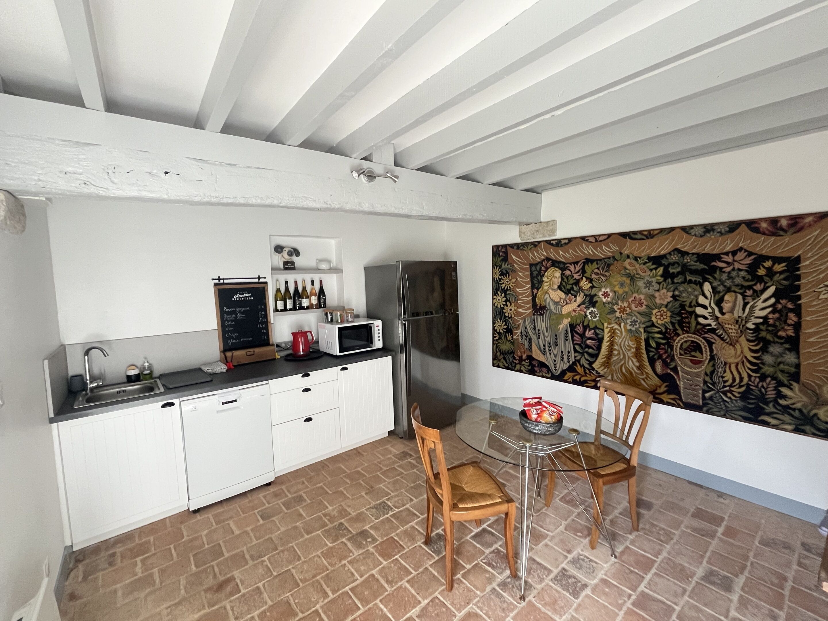 Una cocina con armarios blancos, microondas, hervidor y refrigerador. Hay una mesa con sillas y un tapiz en la pared.