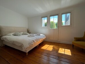 Une chambre avec un plancher en bois, un lit recouvert de draps blancs, un fauteuil jaune et trois fenêtres donnant sur des arbres.