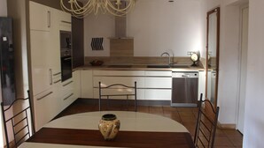 A modern kitchen with white cabinetry, a stainless steel dishwasher, a chandelier, and a dining table with a vase. - House Les Hortensias (MONTAUROUX)