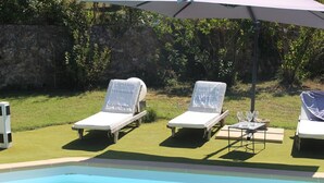 A poolside area with three lounge chairs, a table with two glasses of wine, and an umbrella providing shade. - House Les Hortensias (MONTAUROUX)