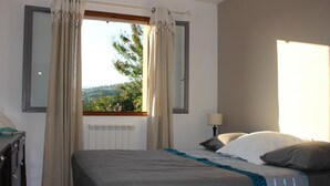 A bedroom with a bed, a dresser, a window with curtains, and a view of greenery outside. - House Les Hortensias (MONTAUROUX)